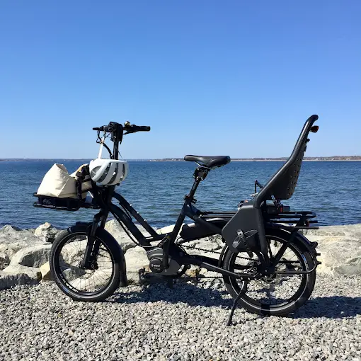 Folding electric bike fitted with a child seat and front cargo bag, parked on a rocky Rhode Island shoreline